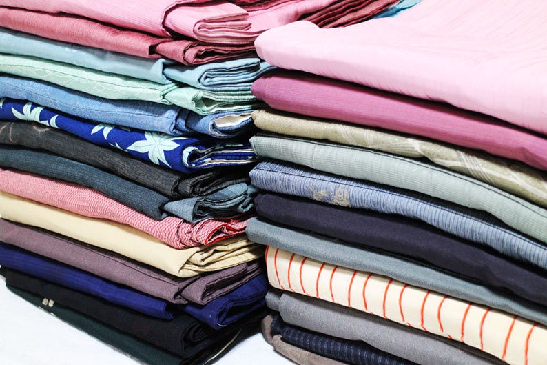 Op de afbeelding: Een stapel gevouwen stof in verschillende kleuren en patronen, waaronder roze, blauw, groen en beige. De stof is netjes gestapeld, met de randen van elk stuk zichtbaar.