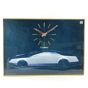 Peut inclure: Horloge murale encadrée avec un design de voiture futuriste. L'horloge a un fond noir et un cadre doré. Les aiguilles et les marqueurs d'heure sont dorés. La voiture est blanche et noire avec des accents bleus. Le texte "AD-I" est visible.