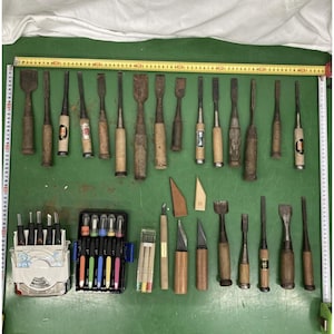 Könnte beinhalten: Eine Sammlung verschiedener Holzbeitel, Schnitzmesser und Präzisionswerkzeuge auf einer grünen Oberfläche. Zwei gelbe Maßbänder rahmen die Werkzeuge ein und zeigen eine Breite von etwa 90 cm und eine Höhe von 60 cm.