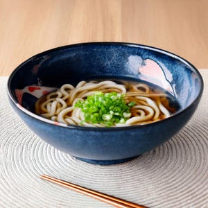May include: A bowl of udon noodles in a dark blue bowl with a fish design. The noodles are topped with chopped green onions and served in a clear broth. A pair of chopsticks rests on the table.