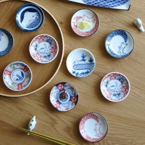 Op de afbeelding: Een verzameling kleine, ronde keramische schaaltjes met diverse handgeschilderde ontwerpen. De schaaltjes hebben blauwe, rode en witte kleuren, met illustraties van dieren en bloemmotieven. Sommige schaaltjes zijn op een houten dienblad geplaatst.