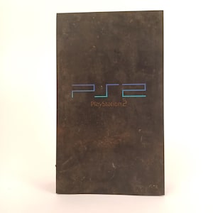 May include: A dark brown PlayStation 2 game case. The front of the case features the stylized letters "PS2" in blue and teal, with "PlayStation 2" printed below. The case has a worn appearance.
