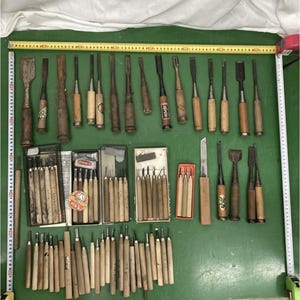Pode incluir: Uma grande coleção de formões para madeira e ferramentas de escultura variadas com cabos de madeira, dispostos sobre uma superfície verde. Duas fitas métricas amarelas emolduram as ferramentas, mostrando 90 cm de largura e 60 cm de altura.