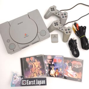 May include: A gray PlayStation console with two matching controllers, a memory card, and three Tekken game cases. Cables and a power cord are also included. The console has the Sony and PlayStation logos.