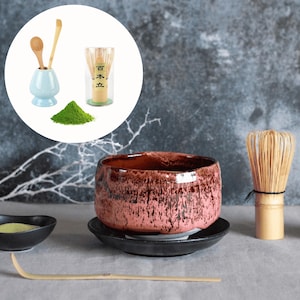 Könnte beinhalten: Ein Matcha-Teeset mit einer rosa-braunen Keramikschale auf einer schwarzen Untertasse, einem Bambusbesen und einem kleinen blauen Keramikhalter mit Holzlöffeln. Grünes Matcha-Pulver und ein Bambuslöffel sind ebenfalls vorhanden.