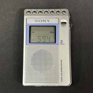 以下が含まれることがあります： 594kHzを表示するデジタルディスプレイを備えたシルバーのソニー製FM/AMラジオICF-R351。上部に7つの円形ボタン、青い枠の画面、円形パターンのスピーカーグリル、右側に小さなライトボタンが配置されています。