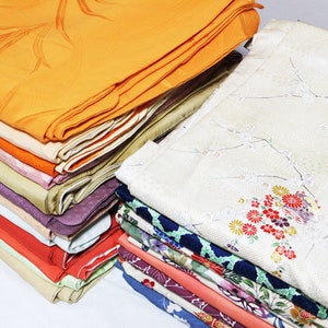 Op de afbeelding: Een stapel gevouwen stof in verschillende kleuren en patronen, waaronder oranje, roze, rood, groen en blauw. De stof is netjes gestapeld, met de bovenste laag dubbelgevouwen.