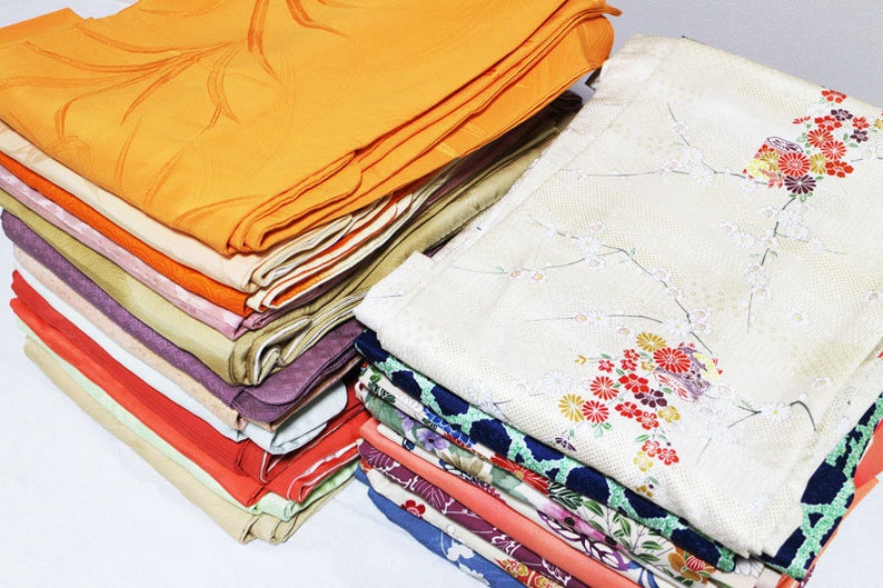 Op de afbeelding: Een stapel gevouwen stof in verschillende kleuren en patronen, waaronder oranje, roze, rood, groen en blauw. De stof is netjes gestapeld, met de bovenste laag dubbelgevouwen.