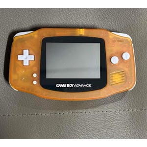 May include: An orange Game Boy Advance handheld console. The device has a black screen, white buttons, and the words "GAME BOY ADVANCE" printed on the bottom. The console is a retro gaming device.