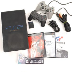 May include: A black PlayStation 2 console with two gray and black controllers, memory card, and game discs. The console has the text "PS2 PlayStation 2" on the front. The game discs include "Gran Turismo 3" and "Gran Turismo 4".
