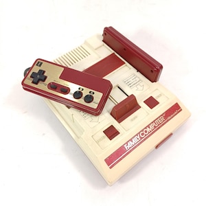 Könnte beinhalten: Eine Vintage Nintendo Family Computer Konsole mit einem Controller. Die Konsole ist cremefarben mit roten Akzenten und dem Schriftzug "FAMILY COMPUTER" auf der Vorderseite. Der Controller ist rot und goldfarben mit schwarzen Knöpfen.