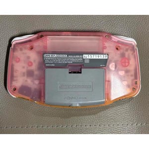 May include: A pink Game Boy Advance console, viewed from the back. The battery compartment and internal components are visible through the translucent pink casing. Model number AGB-001, serial number AJ15719139.