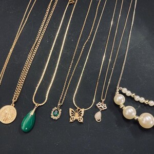 May include: A selection of gold-coloured necklaces with diverse pendants, including an amber-coloured rectangular pendant, a coin, a green teardrop, a butterfly, and a pearl necklace. The necklaces are displayed on a dark surface.