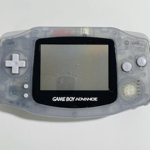 Könnte beinhalten: Eine transparente Game Boy Advance Handheld-Konsole. Das Gerät verfügt über ein Steuerkreuz, A- und B-Tasten sowie einen Power-Knopf. Der Bildschirm zeigt die Worte "GAME BOY ADVANCE" in Weiß an.