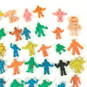 May include: A collection of small, colorful monster figures in various poses. The figures are in shades of orange, pink, green, blue, yellow, and black. Some have horns, while others have unique designs. The toys are likely made of rubber or plastic.
