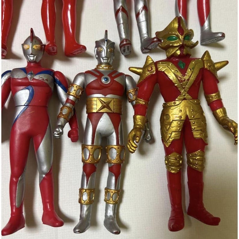 May include: Three standing action figures featuring humanoid designs. The left figure is red and silver with blue accents. The center figure is silver and red with gold circular details. The right figure is red with gold armor plating and green eyes.
