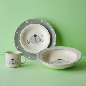 Puede incluir: Un juego de vajilla infantil con un diseño de cara de foca. El juego incluye una taza, un plato pequeño, un plato más grande y un cuenco. Los platos y el cuenco tienen un borde punteado azul claro. La taza, los platos y el cuenco son de color blanquecino.