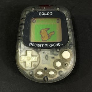 May include: A clear plastic handheld electronic game console with a digital screen displaying a pixelated yellow creature. The device features a white directional pad, three white circular buttons, and two white oval buttons, with the text COLOR and POCKET PIKACHU printed on the front.