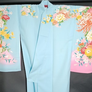 May include: A light blue kimono with floral patterns in shades of pink, yellow, and orange. The kimono features a wrap-around design and is decorated with large, detailed flower arrangements. The sleeves and hem have a pink gradient.
