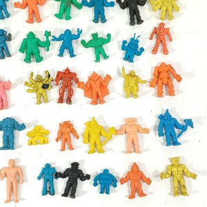 May include: A collection of small, colorful, plastic action figures in various poses. The figures are in shades of orange, green, blue, yellow, and black. Some figures have weapons or unique designs, and they are arranged on a white surface.