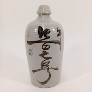 Puede incluir: Una botella de cerámica gris claro con cuello estrecho y cuerpo redondeado. La botella presenta caligrafía marrón oscuro. La botella tiene una forma tradicional, probablemente para almacenar líquidos. La botella es de color neutro.