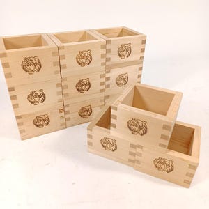 Puede incluir: Un conjunto de tazas de sake de madera clara, cada una con un diseño detallado de cabeza de tigre grabado. Las tazas presentan juntas de cola de milano y vetas de madera visibles, añadiendo un toque rústico.
