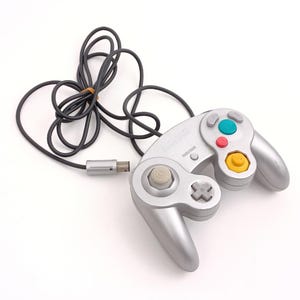 Op de afbeelding: Zilveren videogamecontroller met een zwarte kabel en een USB-connector. De controller heeft een grijs kruis, een witte analoge stick en gekleurde knoppen: groen, rood en geel. De controller staat op een witte achtergrond.