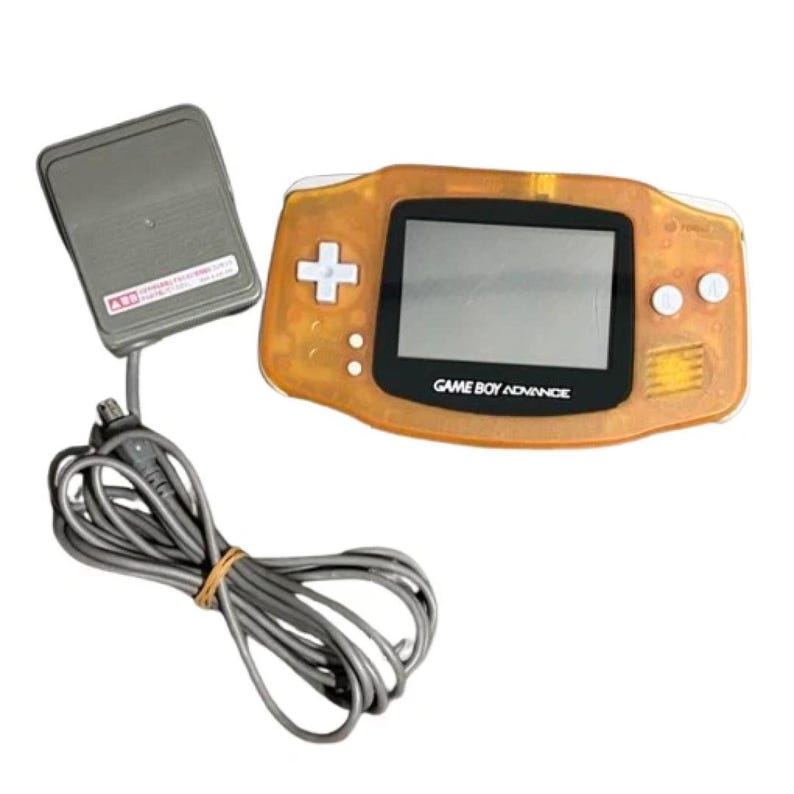 May include: An orange Game Boy Advance handheld console with a black screen and white buttons. The text "GAME BOY ADVANCE" is printed below the screen. A grey power adapter and cable are connected to the console.