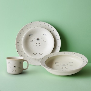 May include: A four-piece off-white dinnerware set with a bear face design. The set includes a plate, a smaller plate, a bowl, and a mug. The plates and bowl have a speckled pattern around the edges. The bear face is drawn with black lines.