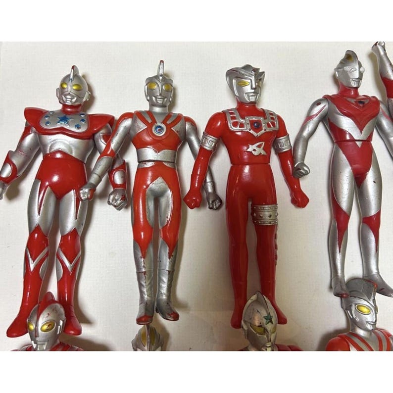 May include: A collection of seven Ultraman action figures featuring red and silver color schemes. Each figure displays distinct chest emblems, head crests, and humanoid proportions. The figures are arranged in a group, showcasing various character designs from the series, with some positioned in the foreground and others standing in the back row.