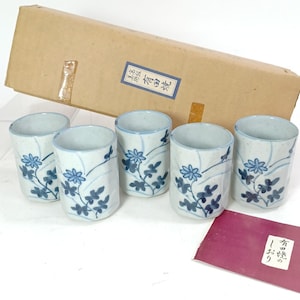 May include: Five ceramic cups with a blue and white floral pattern. The cups are cylindrical with a gently curved rim. The floral design features stylized flowers and leaves. The cups are presented with a cardboard box and a small red card.
