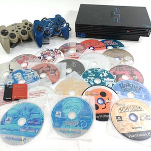 May include: A black PlayStation 2 console, a beige Hori controller, a blue Hori controller, two memory cards, and a collection of various PlayStation 2 game discs in plastic sleeves. The console features the PS2 logo and a disc tray on the front.
