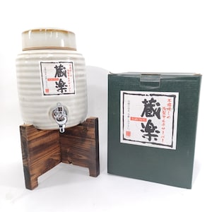 以下が含まれることがあります： 木製の台に置かれた、蛇口付きのセラミック製飲料ディスペンサー。ディスペンサーはオフホワイトで、茶色の蓋と日本語のテキストが書かれたラベルが付いています。同じテキストの緑色の箱が右側にあります。