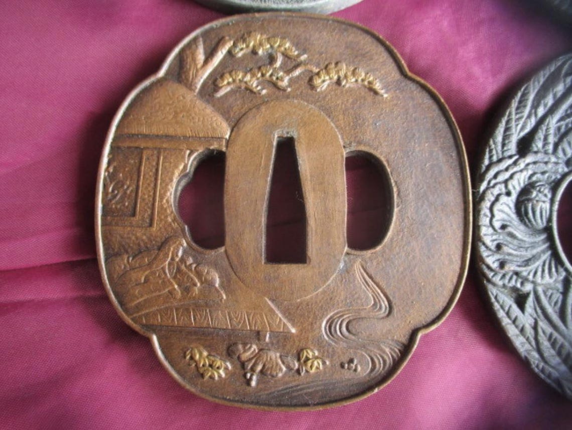 May include: A bronze Japanese tsuba, or sword guard, with a detailed landscape scene. The guard features a central opening and intricate relief carvings of a building, trees, and flowing water. The tsuba has a decorative, scalloped edge.