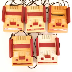 Può includere: Cinque console vintage NES Family Computer con controller rossi e beige. Ogni console ha un pulsante di accensione rosso e uno slot per una cartuccia di gioco.