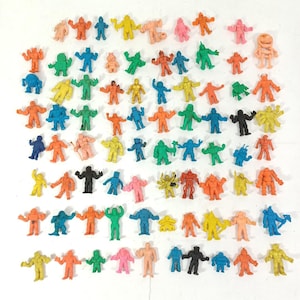 May include: A collection of small, colorful action figure toys in various poses. The figures are in shades of orange, blue, green, yellow, pink, and black. The toys are arranged on a white surface.