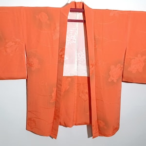 Puede incluir: Un kimono naranja con estampados florales y un interior blanco. El kimono está colgado de una percha. Las mangas son largas y anchas, y la tela parece ligera.