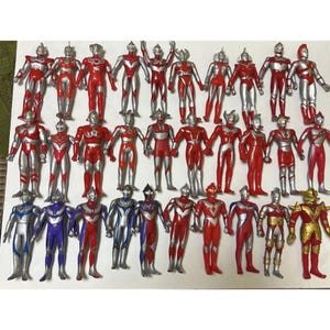 May include: A collection of twenty-seven Ultraman action figures arranged in three rows. The figures are primarily red and silver, with some featuring blue, purple, or gold accents. Each figure stands in a vertical pose with distinct head designs.
