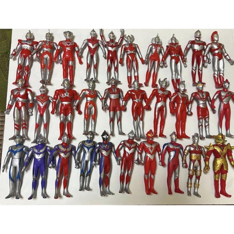 May include: A collection of twenty-seven Ultraman action figures arranged in three rows. The figures are primarily red and silver, with some featuring blue, purple, or gold accents. Each figure stands in a vertical pose with distinct head designs.