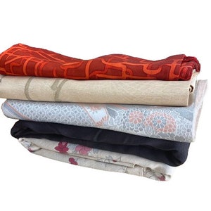 Puede incluir: Una pila de cinco manteles individuales de tela con diferentes patrones. El mantel individual superior es rojo con un diseño geométrico, el segundo es beige con un patrón sutil, el tercero es gris con un patrón floral, el cuarto es negro y el último es beige con un patrón floral rosa.