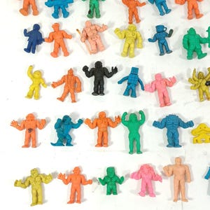 May include: A collection of small, colorful action figure toys in various poses. The figures are in shades of blue, orange, green, yellow, pink, and black. The toys appear to be made of a rubbery material and are arranged against a white background.