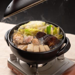May include: A black ceramic pot filled with a steaming hot Japanese hot pot, or nabe, with chicken, tofu, mushrooms, and vegetables. The pot is sitting on a small, silver burner.
