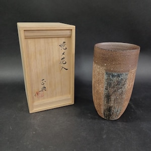 May include: A cylindrical ceramic vase with a textured, mottled brown and dark grey finish, accompanied by a rectangular light-colored wooden storage box featuring black Japanese calligraphy characters and a small red seal on the front panel.
