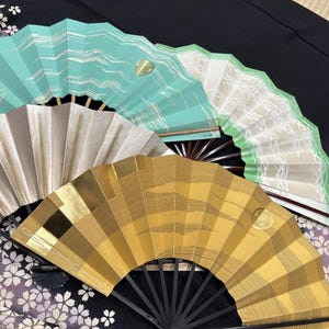 May include: A collection of decorative folding fans in various colors and patterns. The fans include shades of gold, beige, and teal, with geometric and wave-like designs. Each fan has a dark handle and a small, gold emblem.