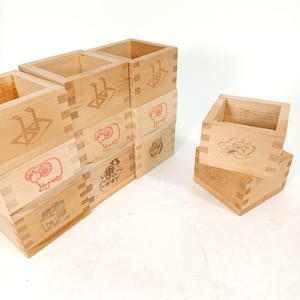 May include: A collection of wooden boxes with dovetail joints. Each box features a different engraved design, including animals and symbols. The boxes are stacked and arranged on a white surface, showcasing their natural wood color and intricate craftsmanship.