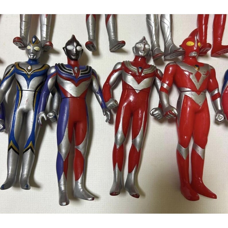 May include: A collection of four standing Ultraman action figures featuring distinct color schemes of silver, blue, and red. Each figure displays a humanoid form with a smooth finish, pointed head crests, and detailed chest emblems, arranged in a row.