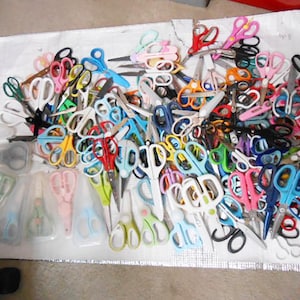 May include: A selection of scissors in various colours, including pink, blue, green, and yellow. The scissors are of different sizes and styles, some with protective covers. The image shows a variety of craft and office scissors.