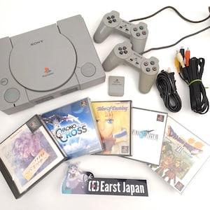 May include: A gray PlayStation console with two matching controllers, a memory card, and several game cases. The game cases include titles like "Chrono Cross" and "Final Fantasy VII". Cables and a power cord are also included.