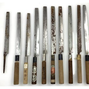以下が含まれることがあります： 長い細身の片刃の金属製ブレードと木製の柄を持つ、日本スタイルの包丁9本のセット。いくつかのブレードには酸化や錆が見られます。一部の柄には、日本語の文字や円形のロゴが印刷された紙のラベルが貼られています。