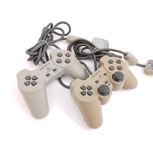 Op de afbeelding: Drie beige Sony PlayStation-controllers met grijze kabels. Elke controller heeft een directioneel pad, actieknoppen en start/select-knoppen. De controllers zijn geplaatst tegen een witte achtergrond.